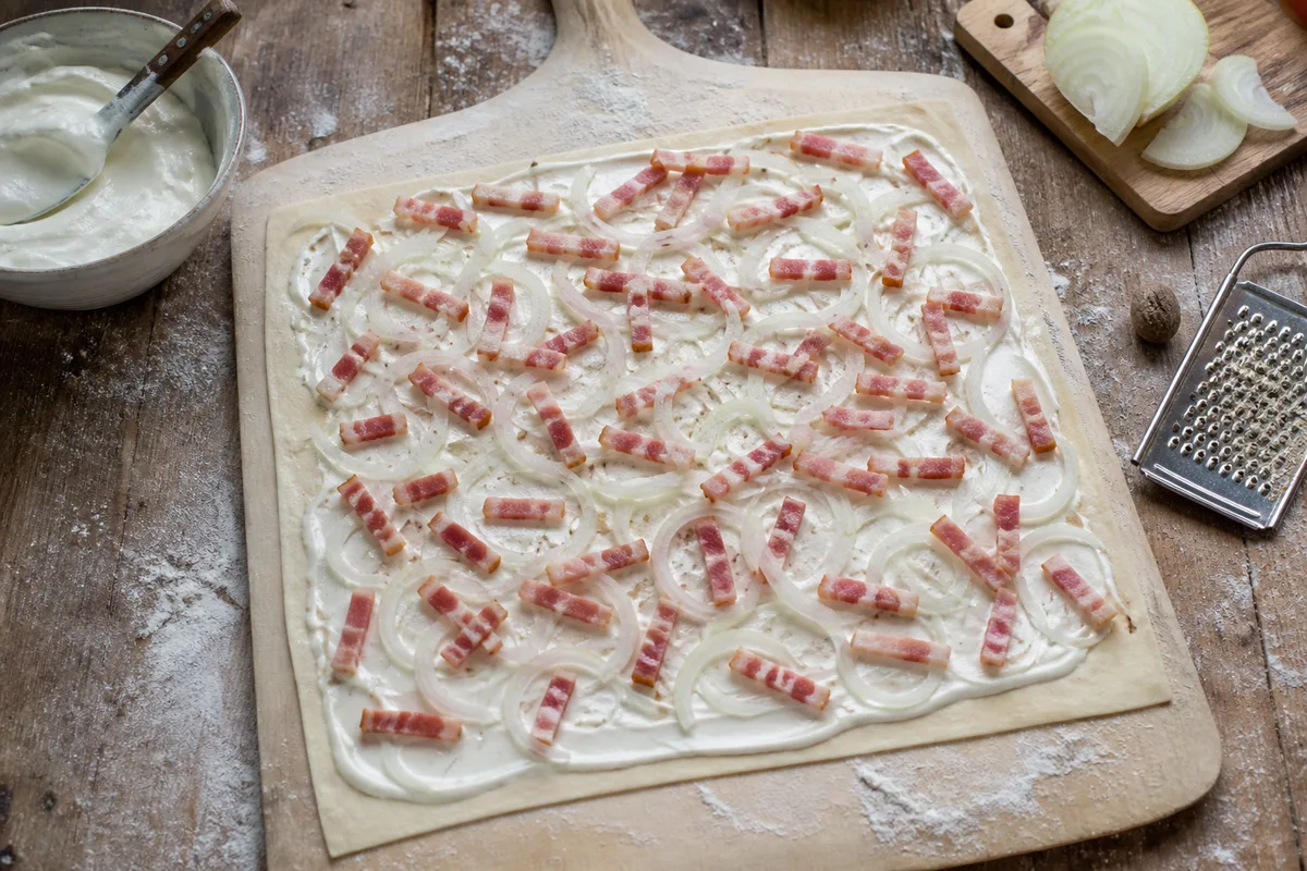 Tarte flambée crue garnie de crème blanche, oignons émincés et lardons fumés sur une pelle en bois